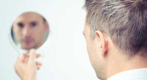 Photo of a man seen from behind looking at his own face in a small hand mirror